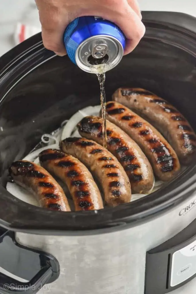 beer being poured over grilled brats with onions underneath in a layer in a slow cooker