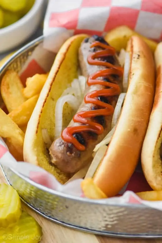 a beer brat in a bun in a tray surrounded by fries garnished with onions and topped with ketchup