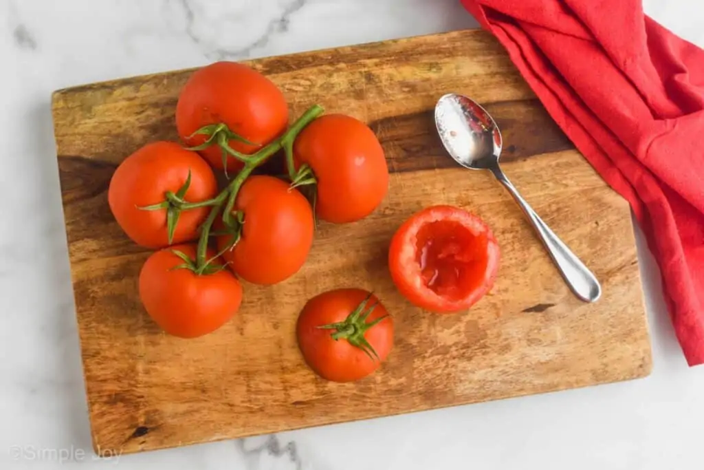 several tomatoes on the vine on a cutting board with one removed, top off, and hallowed out next to a spoon