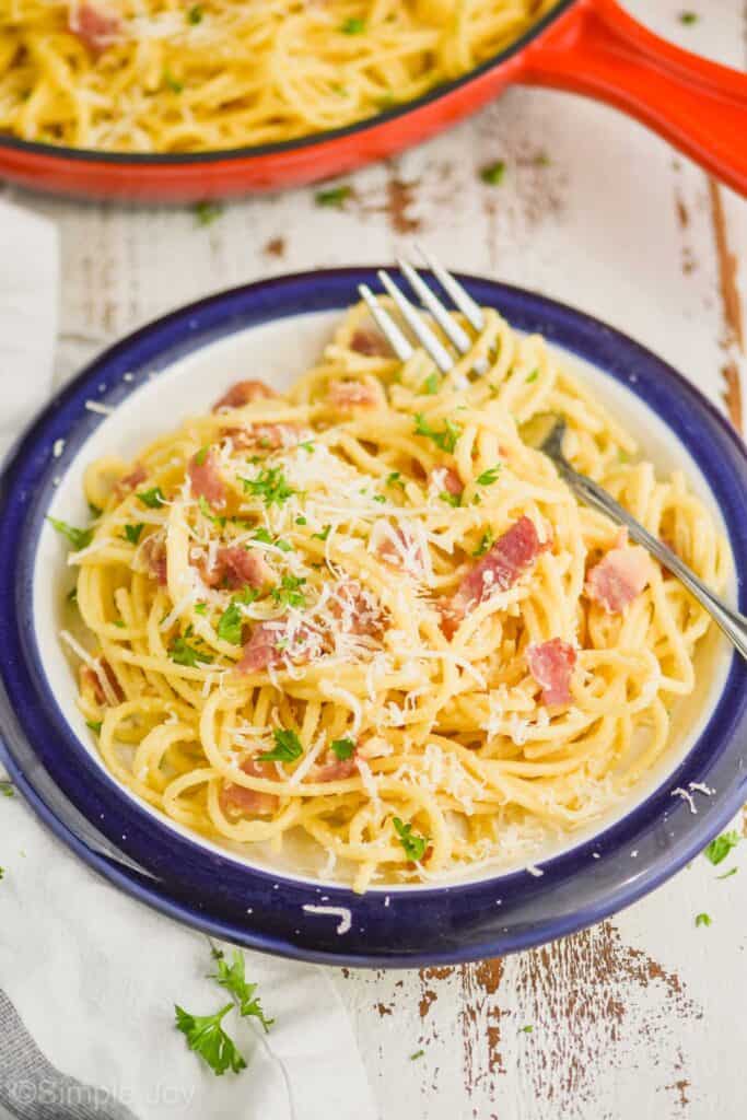 small plate full of pasta carbonara recipe with a fork, garnished with grated parmesan cheese and fresh parsley