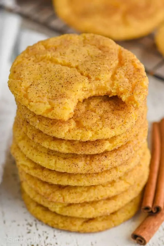 stack of pumpkin snickerdoodle cookies with the top one missing a bite, two cinnamon sticks sitting next to it