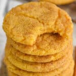 stack of pumpkin snickerdoodle cookies with the top one missing a bite, two cinnamon sticks sitting next to it