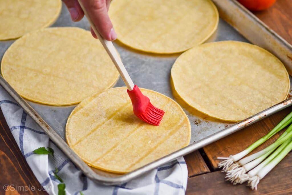 a hand using a silicon brush to spread vegetable oil onto an uncooked corn tortilla on a rimmed baking sheet with five other corn tortillas