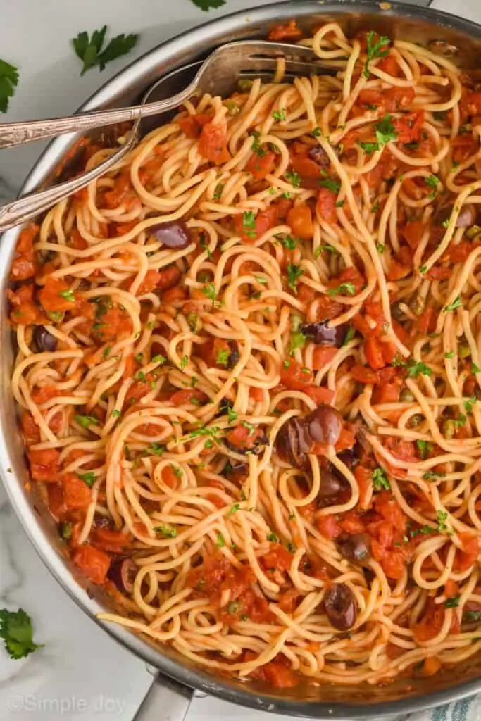 overhead view of pasta puttanesca in a skillet with two serving utensils