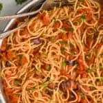 overhead view of pasta puttanesca in a skillet with two serving utensils