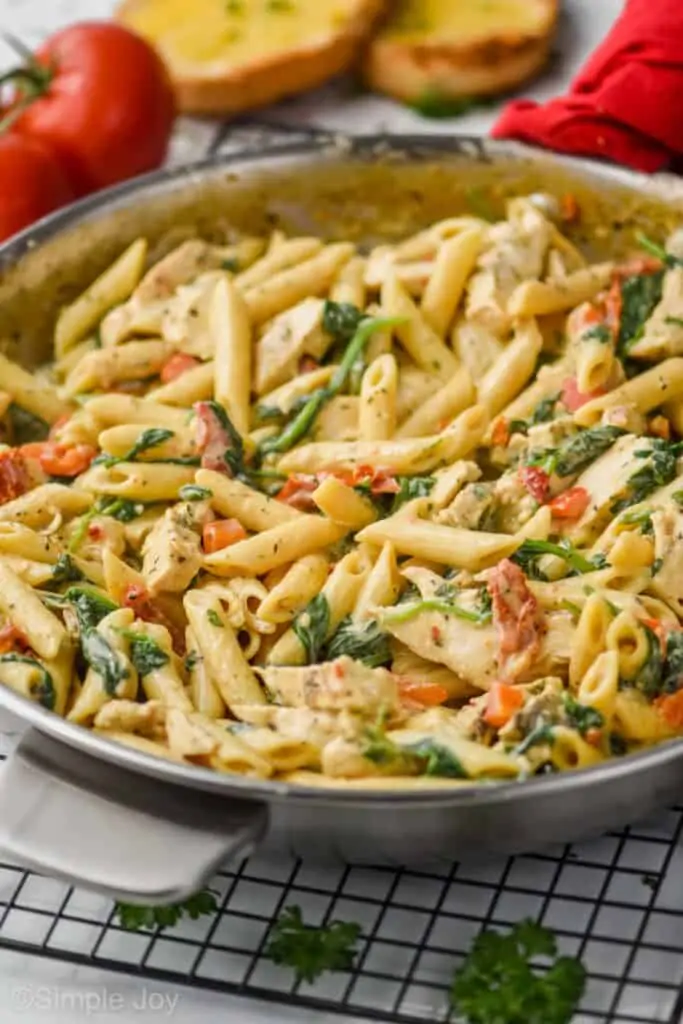 side view of a pan of creamy tuscan chicken pasta in a skillet with tomatoes and toasted garlic bread in the background