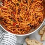 Pinterest graphic for Pasta Puttanesca. Image shows overhead of skillet of Pasta Puttanesca with sliced baguette sitting beside. Says “25 minute Puttanesca recipe simplejoy.com"