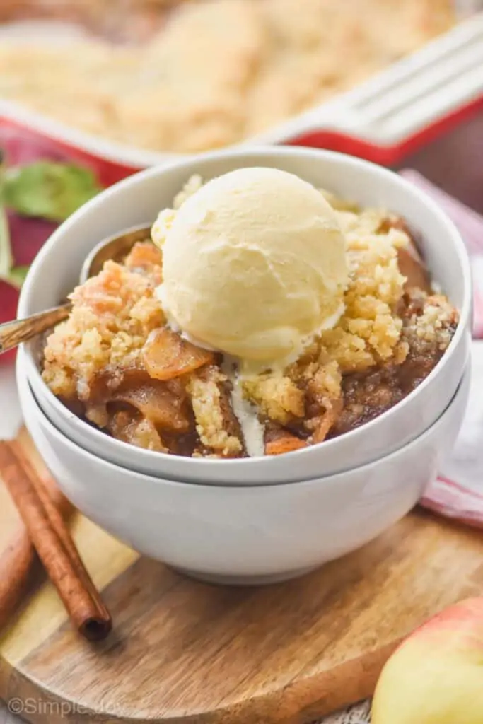 two white bowls stacked holding best apple crisp recipe topped with ice cream, baking dish in the background