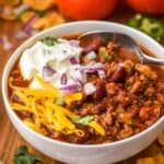 close up of a white bowl full of crockpot chili topped with sour cream, shredded cheese, sour cream, diced onion, fresh cilantro