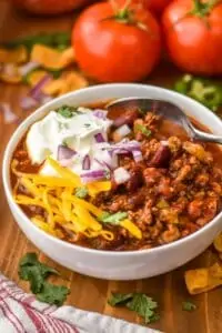 close up of a white bowl full of crockpot chili topped with sour cream, shredded cheese, sour cream, diced onion, fresh cilantro