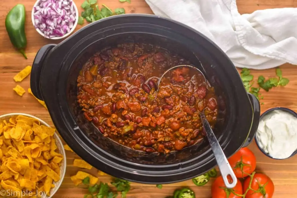 overhead view of crockpot chili recipe in a slow cooker surrounded by chips, onions, tomatoes, and sour cream for toppings