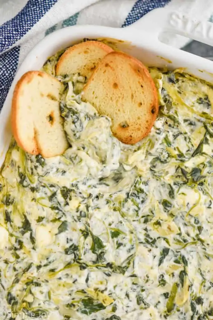 close up of hot spinach artichoke dip in a white casserole dish with bagel chips dipped in