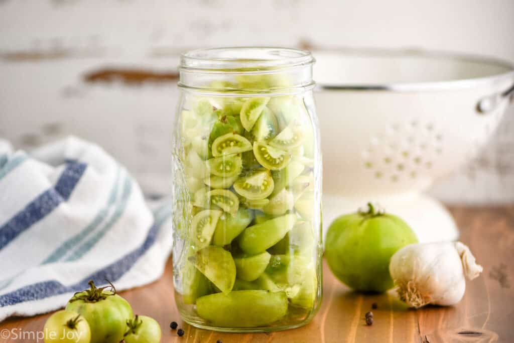 landscape photo of a jar of slices cut up green tomatoes waiting to be pickled next to a garlic bulb