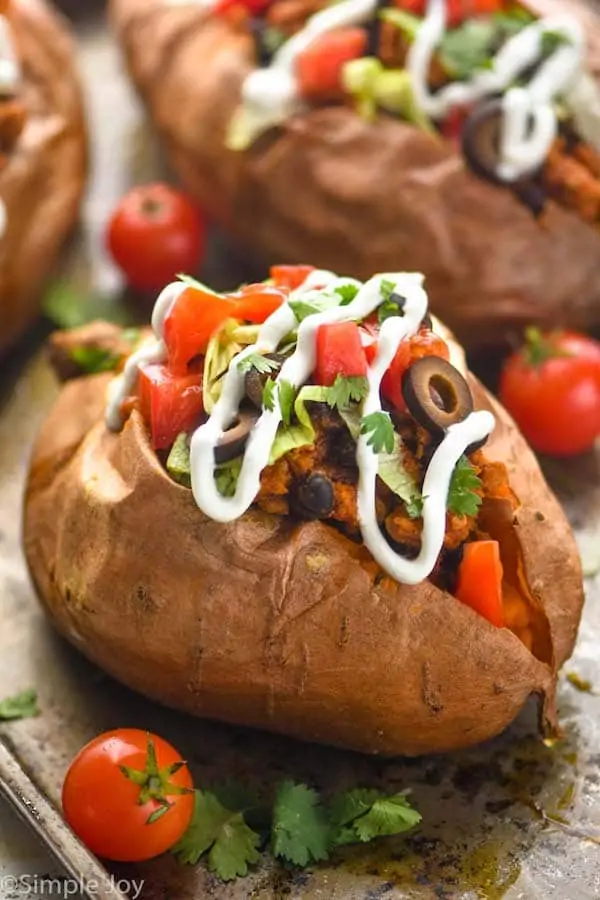 close up of a taco stuffed sweet potato on a baking sheet that has been topped with sour cream, cilantro, sliced olives, diced tomatoes, and lettuce