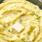 side view of a bowl of creamy mashed potatoes with a pad of melting butter, parsley, and black pepper