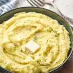 side view of a bowl of creamy mashed potatoes with a pad of melting butter, parsley, and black pepper