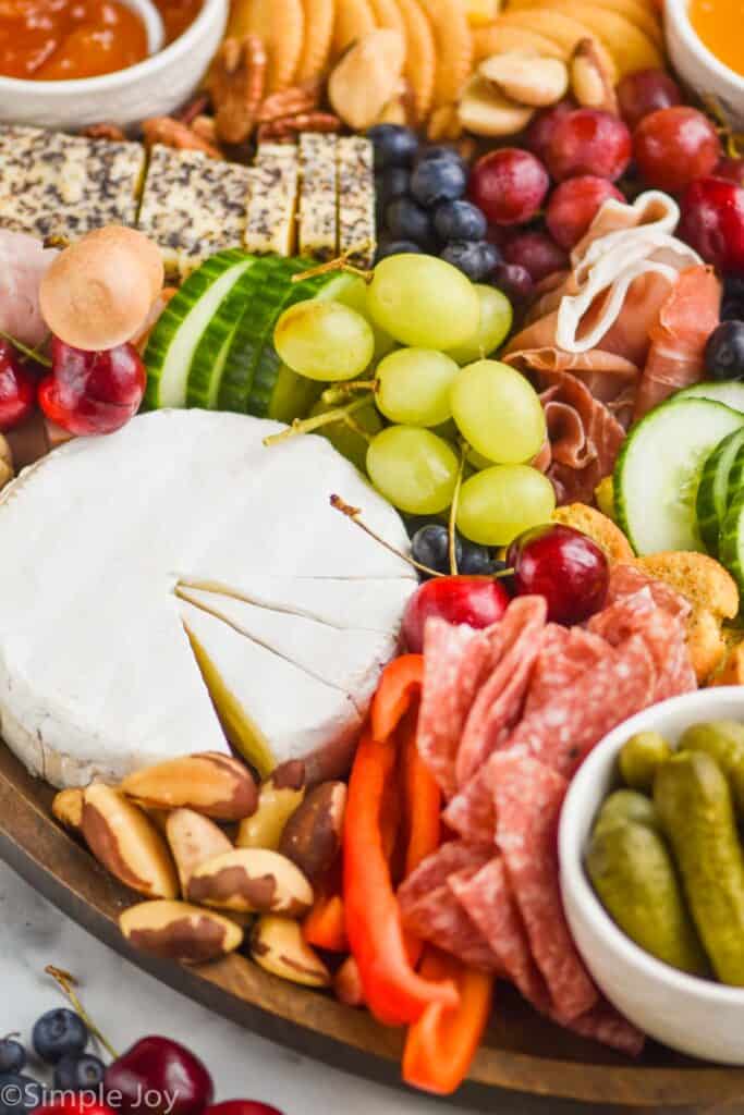 a close up view of a block of brie cut into wedges on a charcuterie board with grapes, meats, cucumbers, berries, and crackers