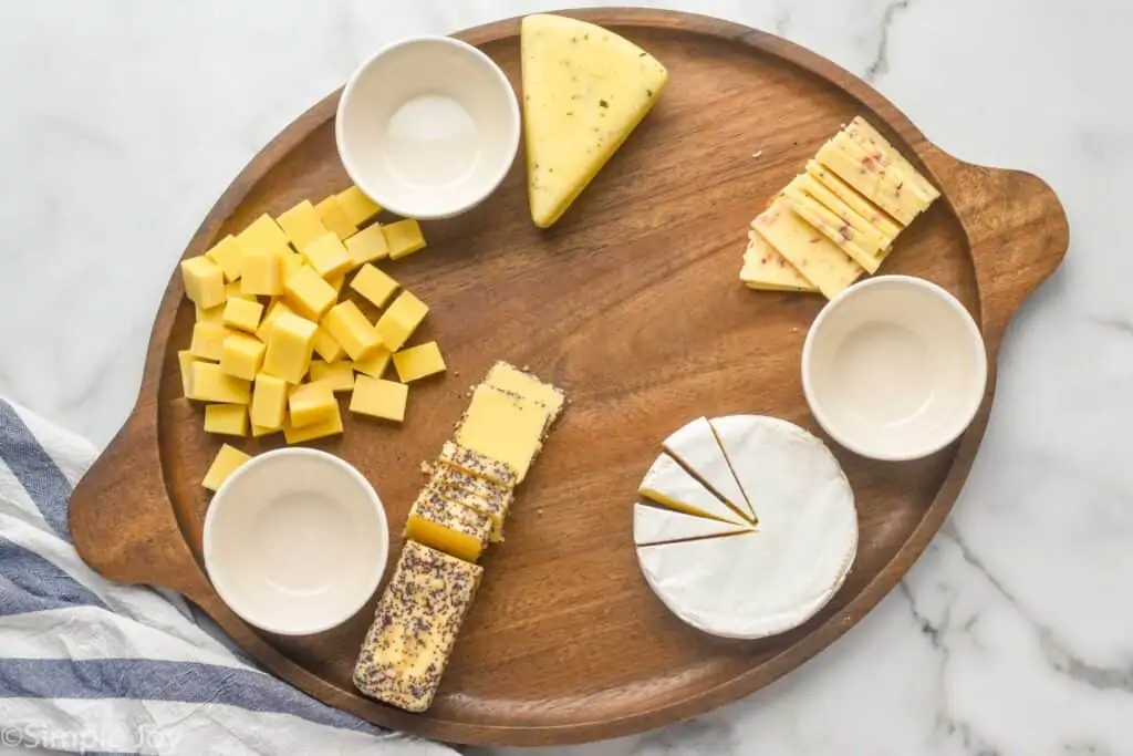 a large wooden tray with three white bowls for condiments and cut up cheeses
