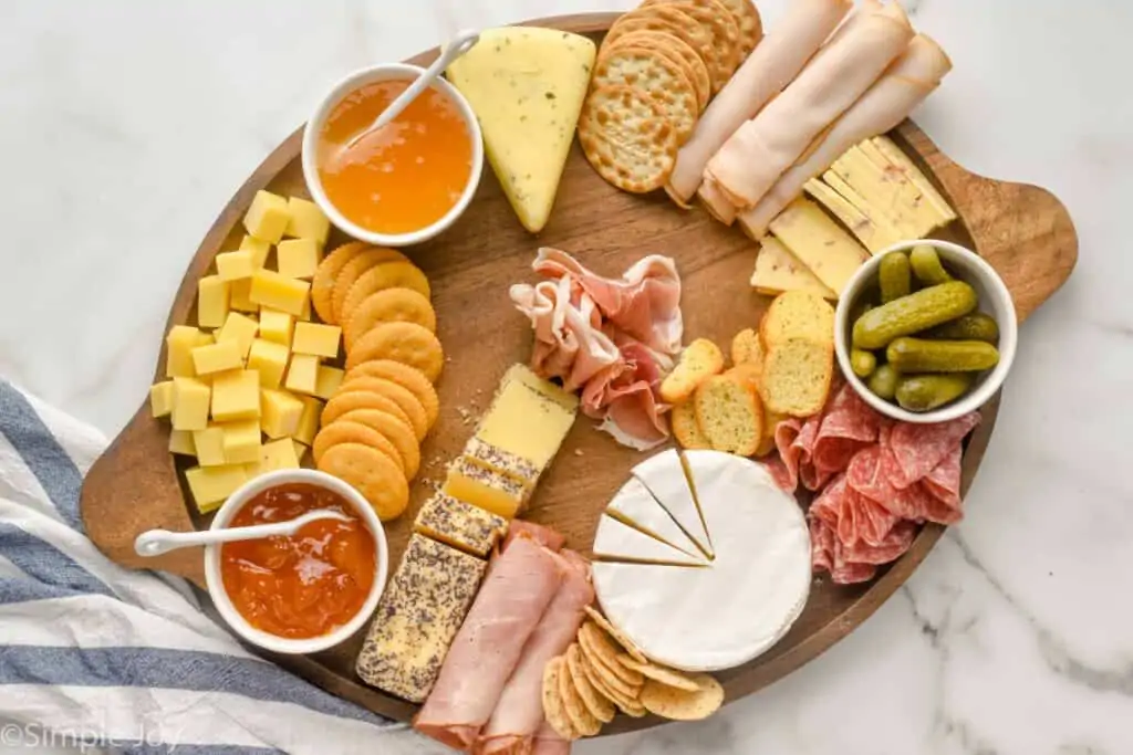 a large wooden tray with bowls of condiments, cut up cheese, deli meat, and crackers