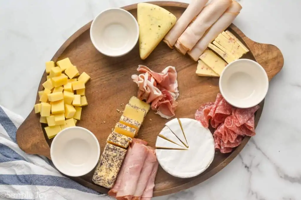 a large wooden tray with three white bowls, deli meats, and four blocks of cheese cut