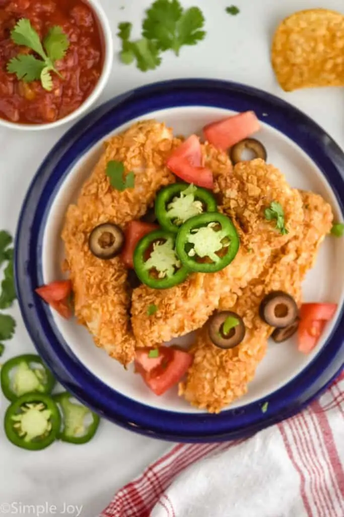 overhead view of a plate of chicken tenders topped with taco toppings like jalapeños, tomatoes, olives, and cilantro