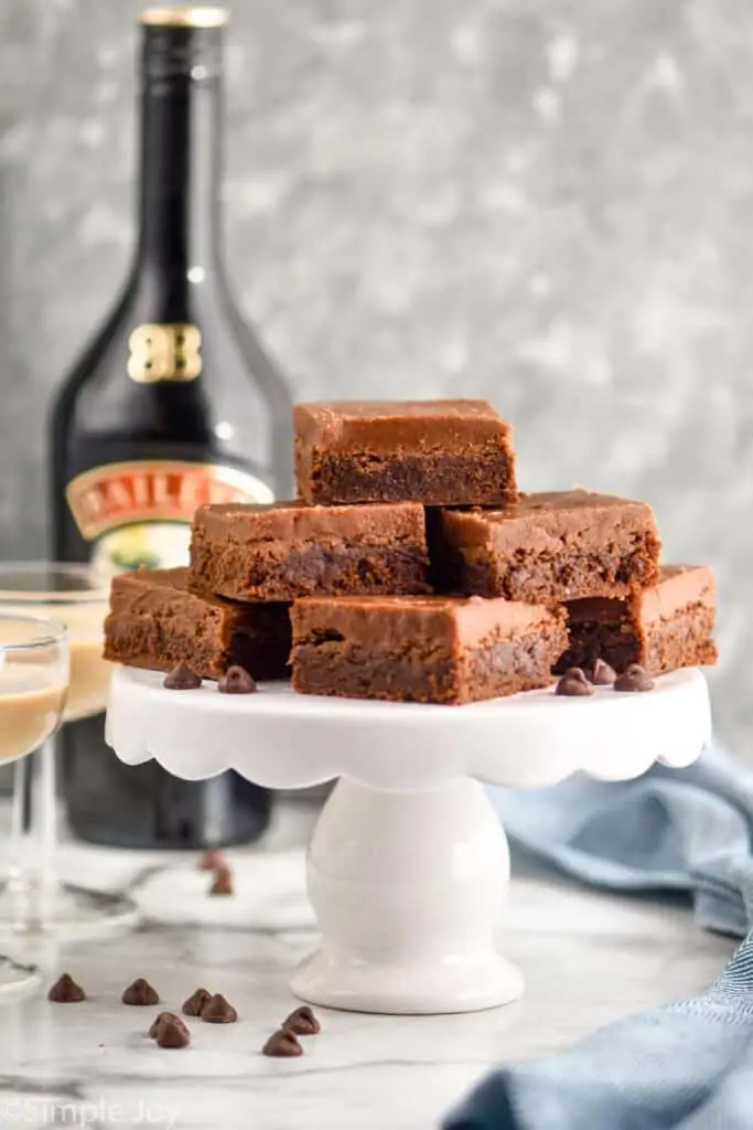 a pile of brownies on a white cake platter with baileys Irish cream in the background
