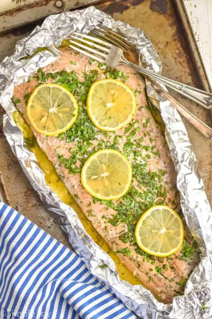 overhead view of baked salmon recipe that is in foil and topped with lemon slices fresh parsley and has two forks next to it