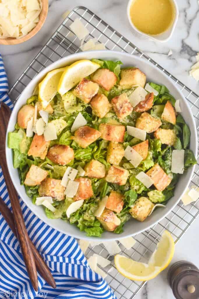 overhead view of a bowl of Caesar salad with dressing next to it, lemon wedges, and additional parmesan peels