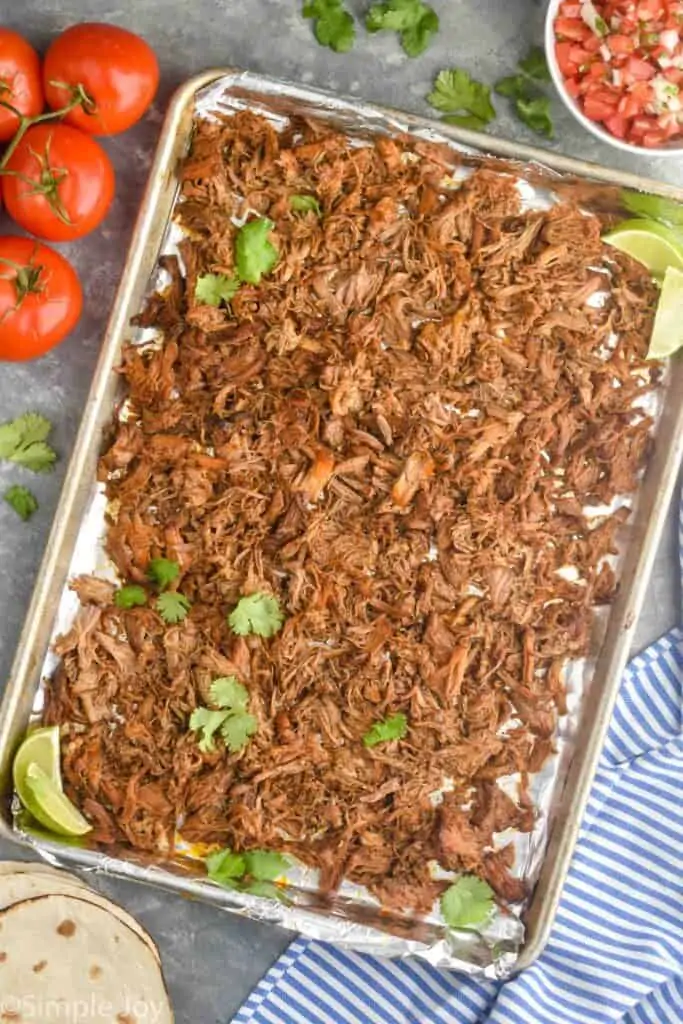 baking sheet full of the pulled meat from a carnitas recipe topped with fresh cilantro and two limes