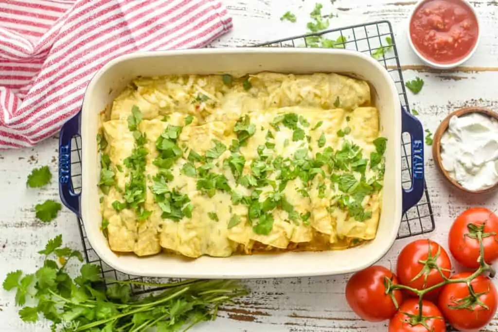 overhead photo of green enchiladas in a casserole dish next to cilantro, a bowl fo salsa, a bowl of sour cream, tomatoes on a vine