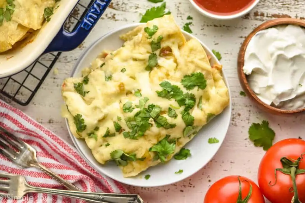 overhead photo of three green chicken enchiladas on a plate with a bowl of sour cream next to them.