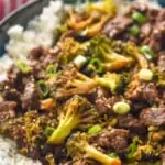 side view of a bowl of beef and broccoli over rice garnished with sesame seeds and sliced scallions