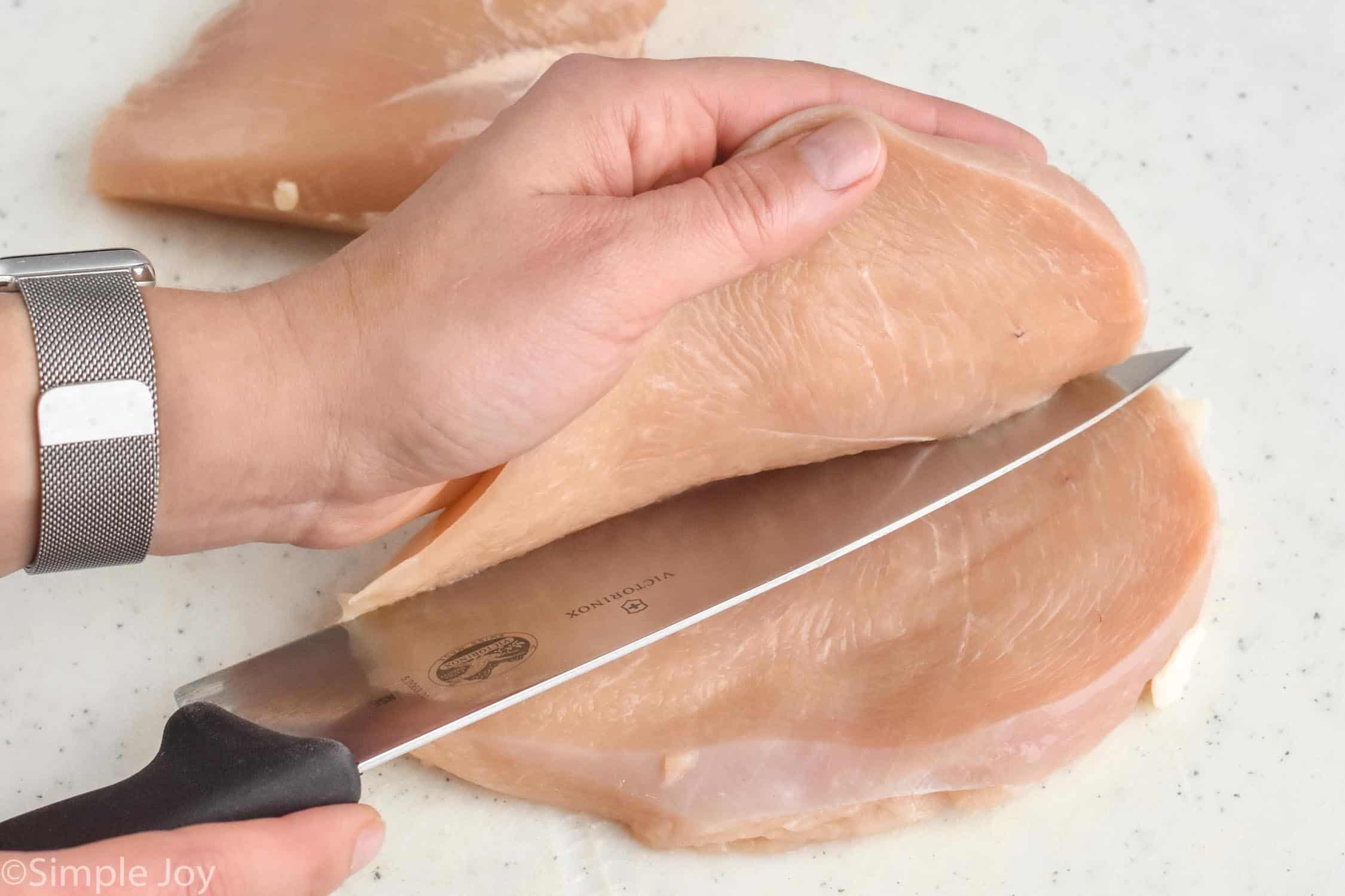a hand pulling back the top half a chicken breast as a knife cuts it in half for chicken cutlets