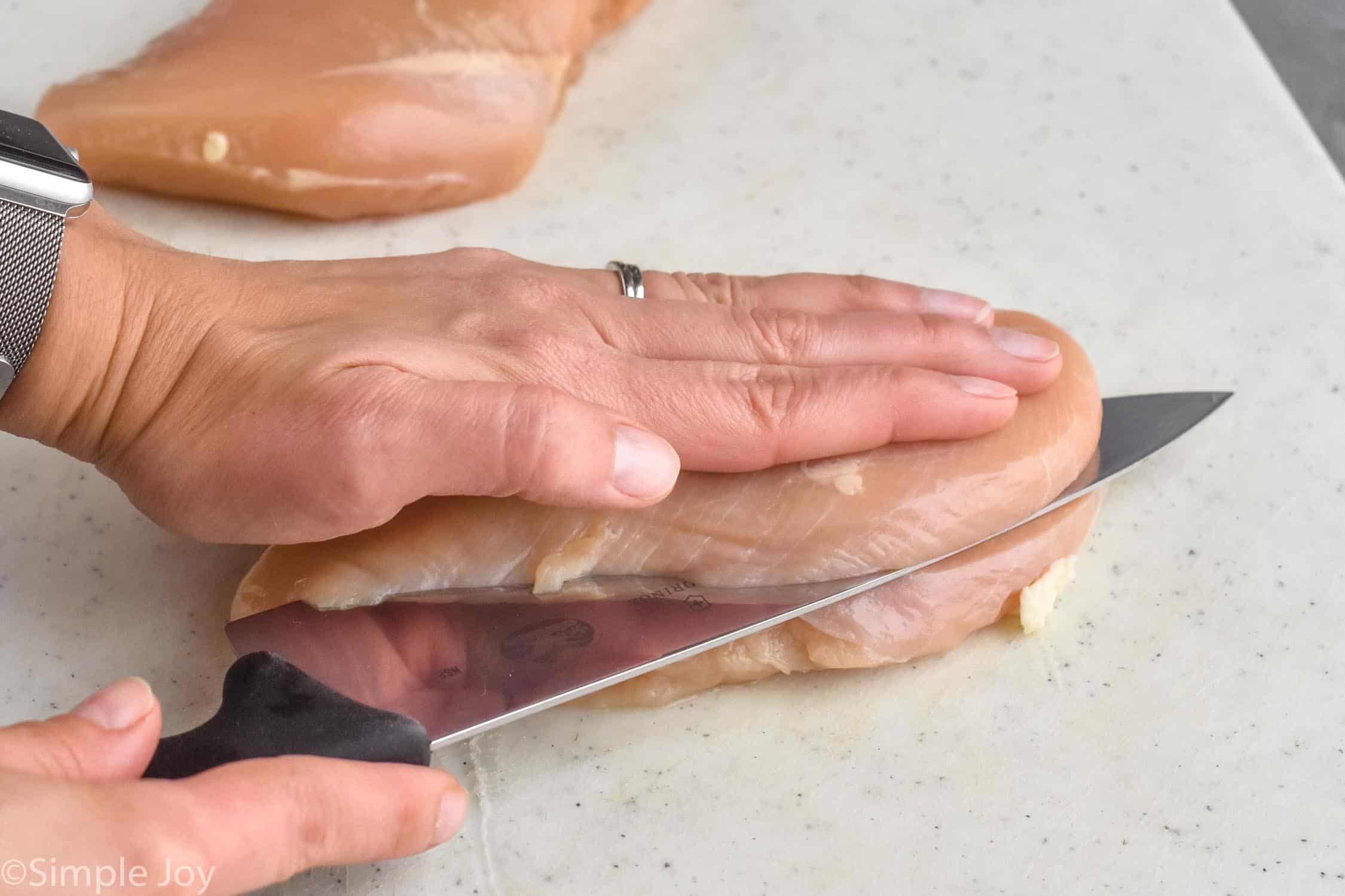 a hand on top of a chicken breast as a knife cuts it in half to make chicken cutlets