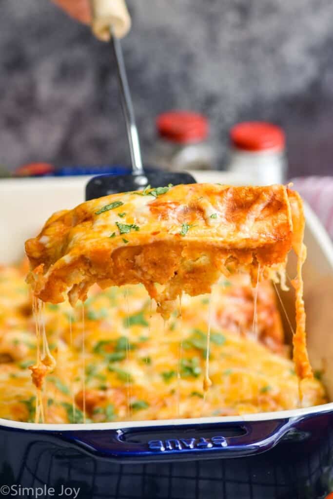 a single enchilada being lifted out of a casserole dish full of chicken enchilada recipe