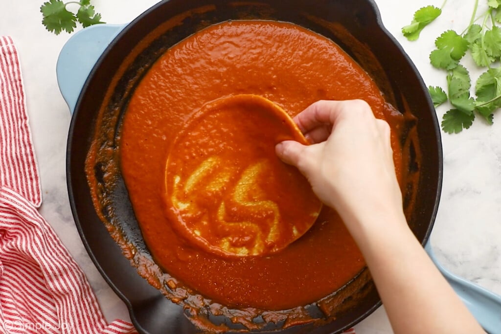 Overhead of skillet of enchilada sauce with person's hand holding a corn tortilla in the sauce to make chicken enchiladas - 8