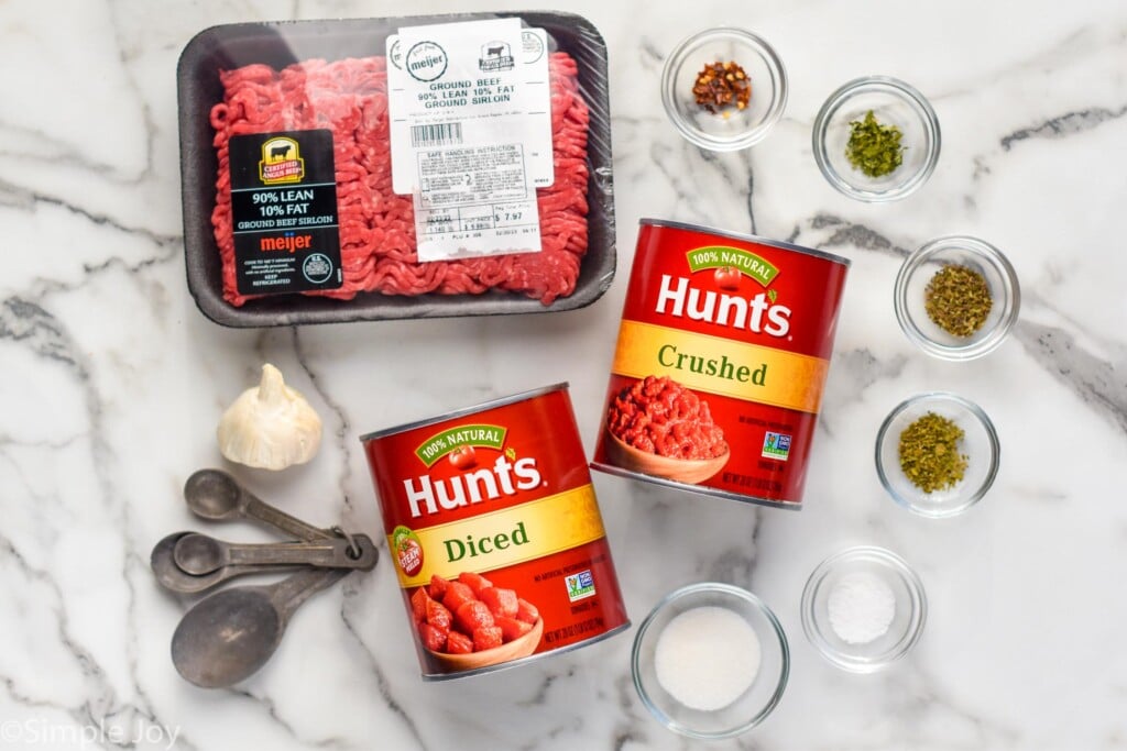 overhead photo of ground beef, spices in small bowls, garlic, and two cans of tomatoes to make meat sauce recipe