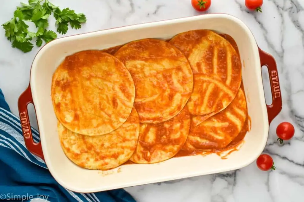 overhead of a baking dish showing six corn tortillas coated in enchilada sauce in a bottom layer