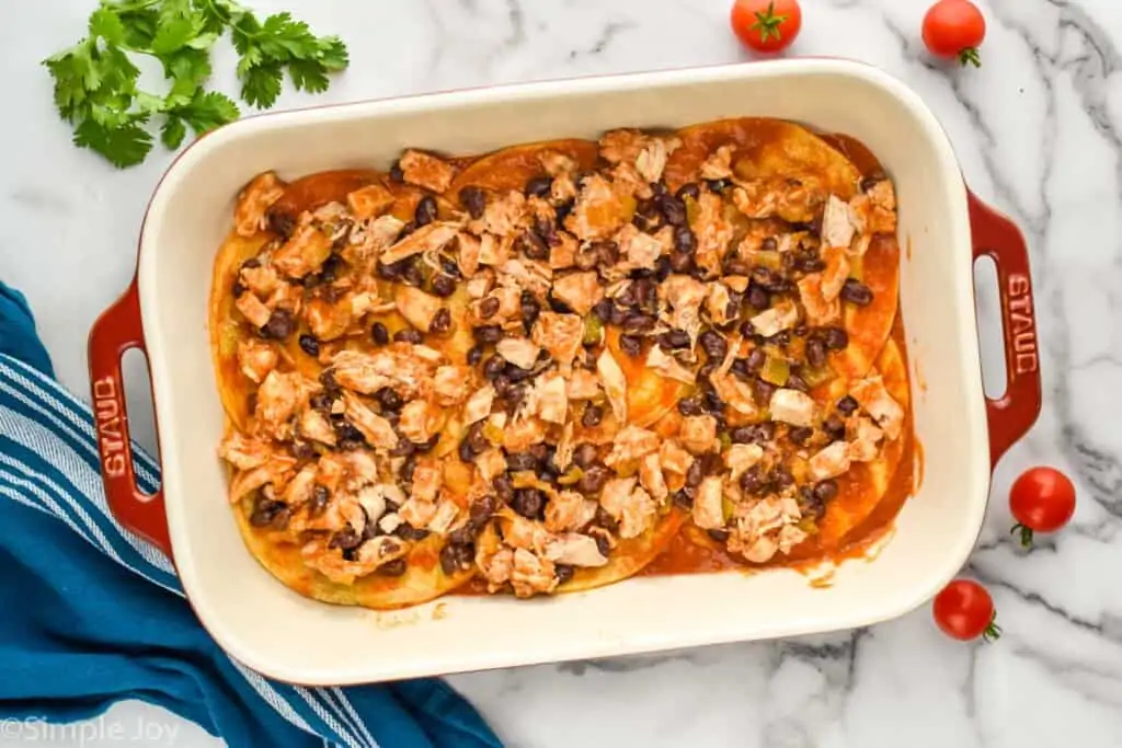 overhead of a baking dish showing layering of casserole, black beans and chicken over enchilada sauce covered tortillas