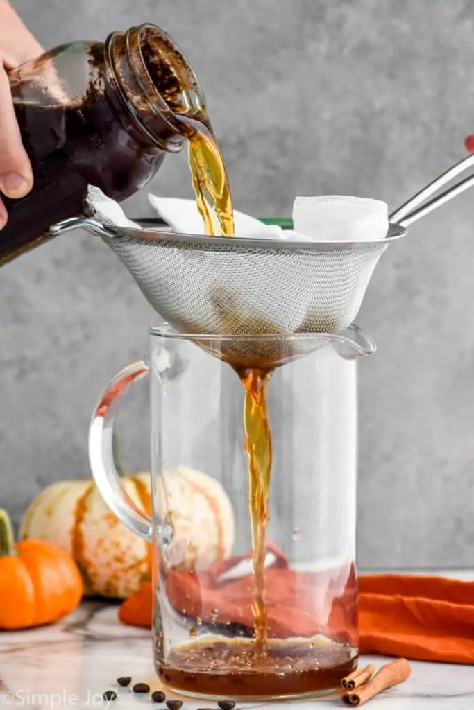 straining pumpkin cold brew with a metal strainer and a cheesecloth into a pitcher