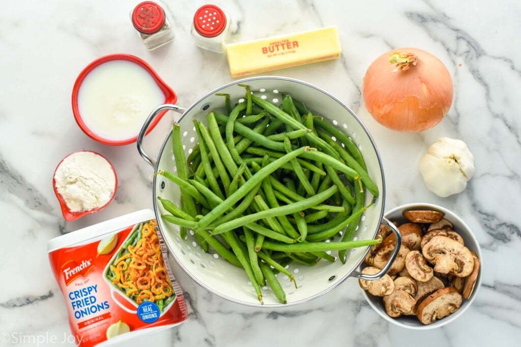 overhead photo of green beans in a colander, mushrooms, garlic, onion, butter, salt and pepper, milk, flour, and crispy fried onions - all the ingredients for green bean casserole from scratch