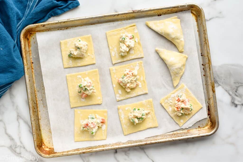 overhead of puff pastry squares on a baking sheet with chicken pot pie filling on them and two folded into triangles