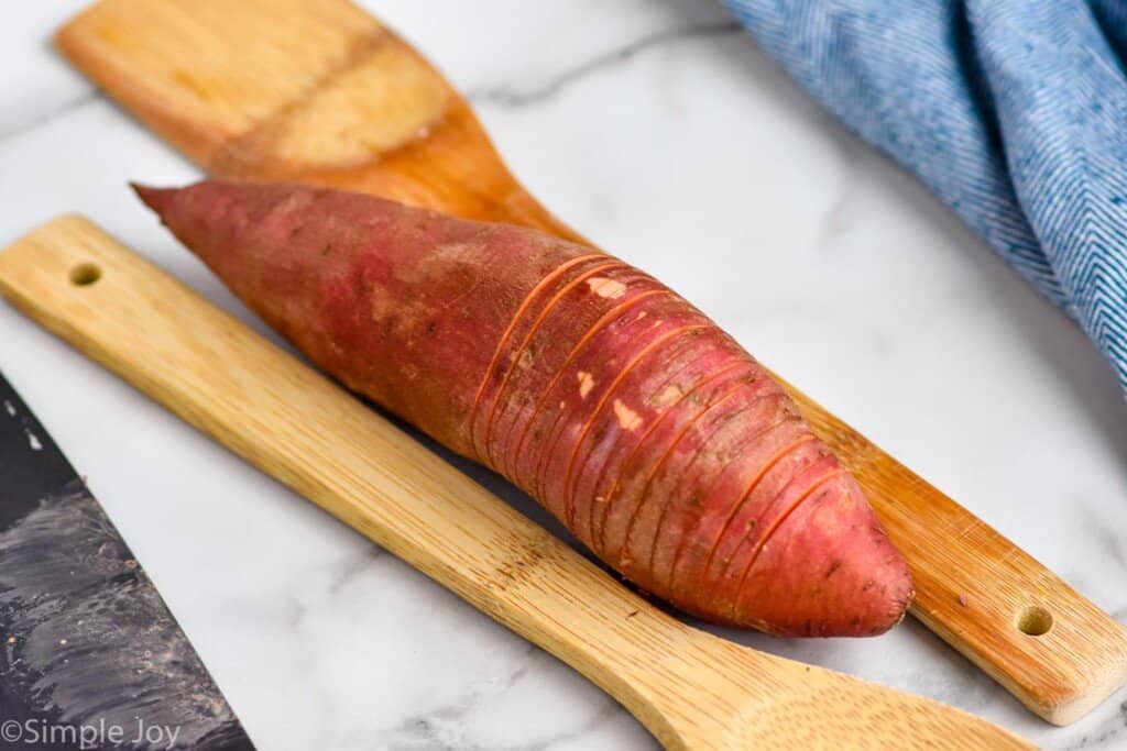 a sweet potato with two wooden spoons, one on each side, that is being cut hasselback style