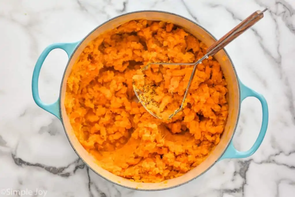 overhead of a dutch oven filled with a mashed sweet potatoes and a potato masher