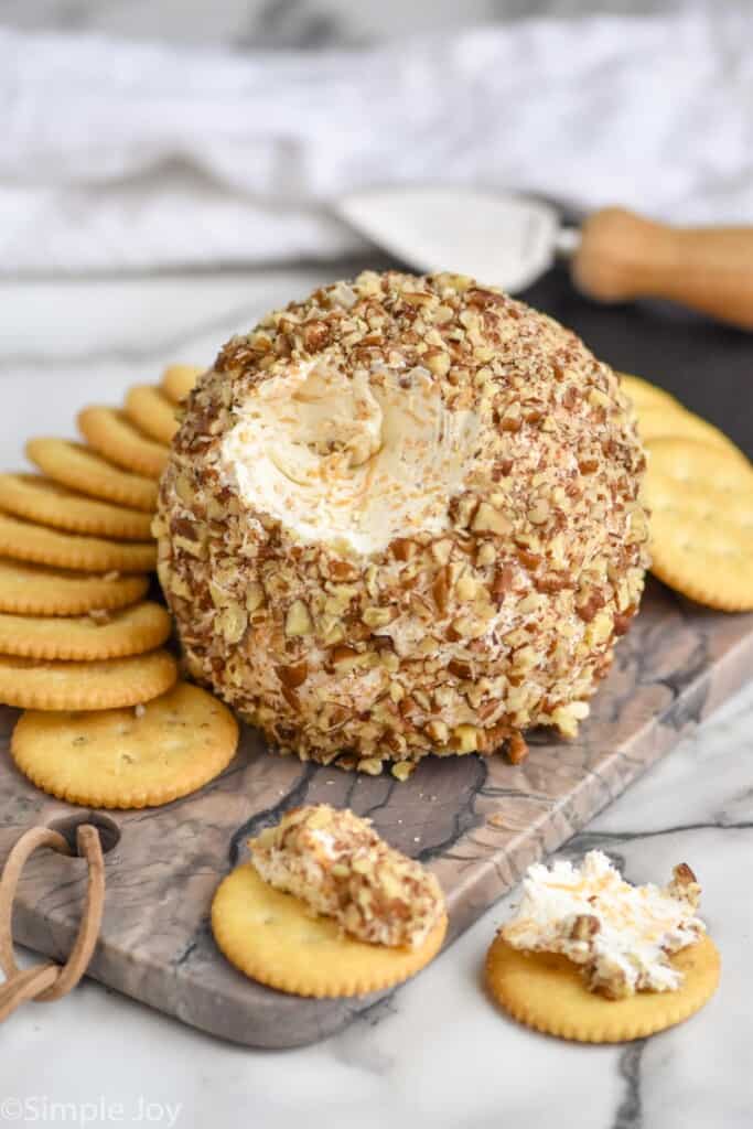 a cheese ball on a marble cutting board with a bit missing, surrounded by crackers and a cheese spreader in the background