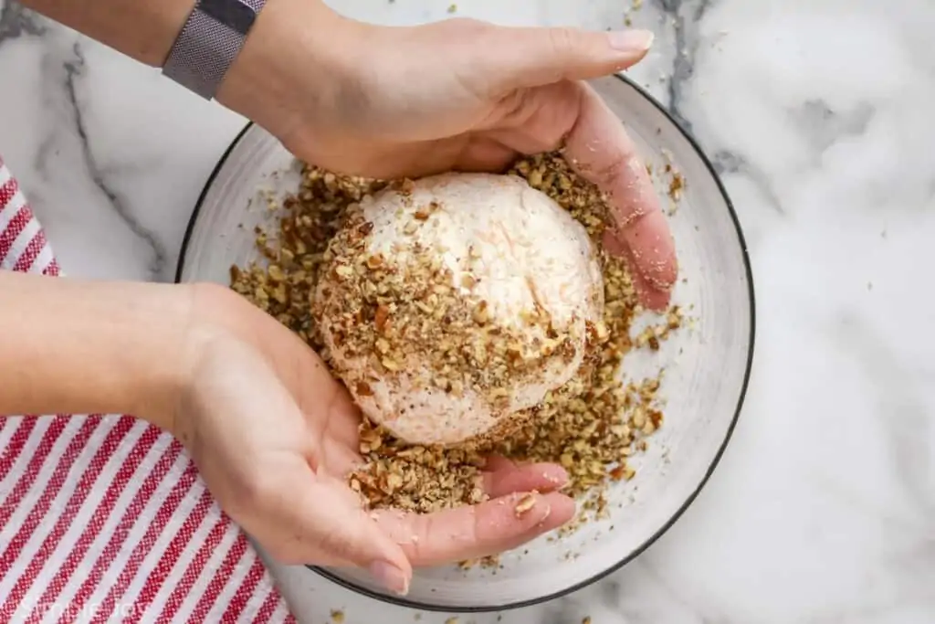 overhead of a cheeseball with hands putting chopped pecans on the outside