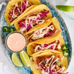 overhead of five fish tacos on a plate next to a small jar of fish taco sauce