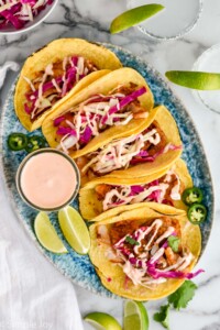 overhead of five fish tacos on a plate next to a small jar of fish taco sauce