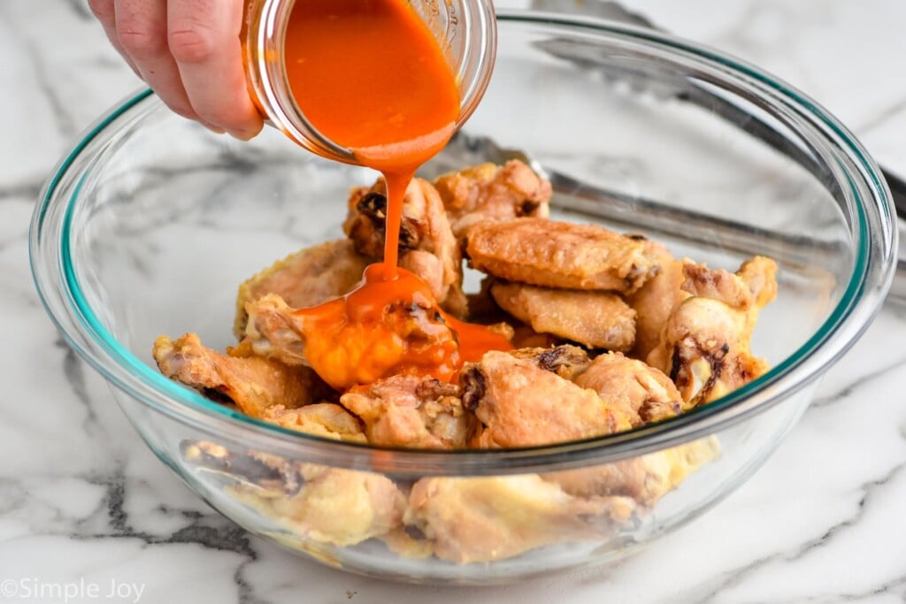 pouring buffalo sauce over a bowl full of crispy chicken wings that have been baked in the oven