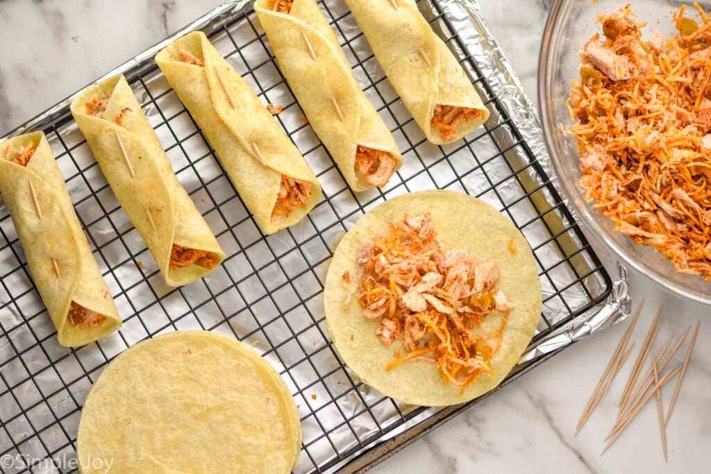 overhead of a baking sheet fit with a wire rack, chicken taquitos in the process of being rolled , being held together with toothpicks